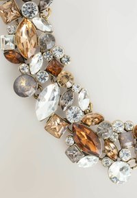 Collana in tono oro con varie gemme tagliate in sfumature di marrone, trasparente e blu, con una combinazione di forme rotonde e geometriche.