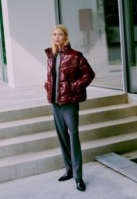 Burgundska sjajna puf jakna s visokim ovratnikom, nosi se preko sive veste. Uparena sa sivim hlačama i crnim čizmama do gležnja, prikazana na otvorenom.