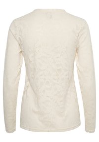 Långärmad ljuskrämfärgad spetsblus med ett blommönster, rund halsringning och en texturerad yta med vågiga manschetter och nederkant.