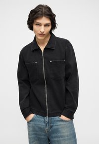 Νεαρό άτομο με μεσαίου μήκους σκούρα μαλλιά φοράει μαύρο μπουφάν με φερμουάρ και μπροστινές τσέπες, μπλε τζιν, με τα χέρια στις τσέπες, ουδέτερο φόντο.