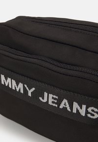 Černý látkový sáček se dvěma paralelními zipy a vodorovným černým popruhem, na kterém je v bílém písmu zobrazeno jméno značky "Tommy Jeans".
