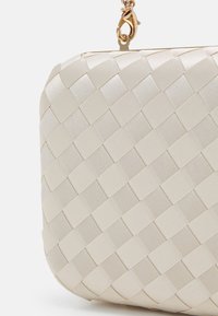 Gros plan d'une pochette tissée couleur crème avec un coin arrondi et un fermoir chaîne dorée en haut sur fond blanc.
