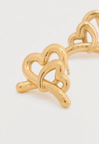 Σκουλαρίκι από μέταλλο σε χρυσαφί απόχρωση με φυλαχτά σε σχήμα καρδιάς που είναι πλεγμένα μεταξύ τους, με λείπασμα λείου φινιρίσματος και χαραγμένη την επιγραφή «GUESS» στη μία πλευρά.