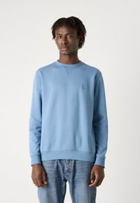 Sudadera de color azul claro con cuello redondo, puños y cintura de canalé. Presenta un pequeño parche de logo rectangular en el pecho. Textura suave.