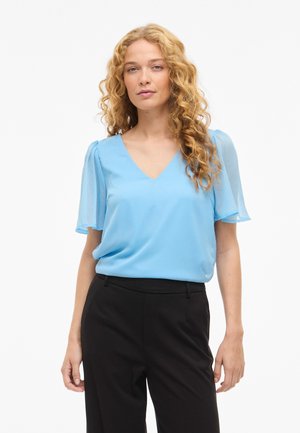 Hosszan göndör szőke hajú nő világoskék V-nyakú blúzt visel áttetsző rövid ujjakkal és fekete magas derekú nadrágot, fehér háttér előtt áll.