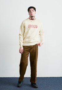 Κρέμα φούτερ με την ένδειξη "AIR" σε κόκκινα γράμματα, σε συνδυασμό με καφέ παντελόνι. Διαθέτει ραμμένες μανσέτες και χαλαρή εφαρμογή. Μαύρα αθλητικά παπούτσια ολοκληρώνουν την εμφάνιση.