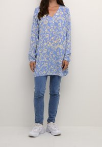 Femme portant un tunique à manches longues florale bleue, un jean skinny bleu et des baskets blanches, se tenant devant un fond clair uni.