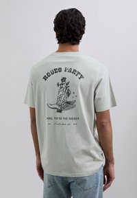 Mann mit dunklen lockigen Haaren, der ein hellgraues T-Shirt mit einem schwarzen "Rodeo Party"-Grafik und Text auf dem Rücken trägt, kombiniert mit hellblauen Jeans.