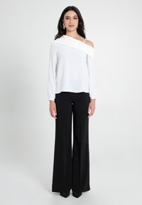 Blusa bianca, scollata su una spalla, con maniche lunghe abbinata a pantaloni neri a gamba larga e tacchi a punta. Tessuto liscio con un design minimalista.
