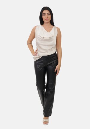 Donna con lunghi capelli neri che indossa un top bianco senza maniche drappeggiato, pantaloni neri in pelle e tacchi bianchi open toe, in piedi con una mano sul fianco.