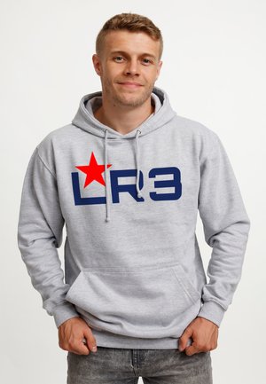 Ung mand smilende, iført en lysegrå hættetrøje med en rød stjerne og navyblåt "LR3"-tekst, poserende foran en ensartet hvid baggrund.