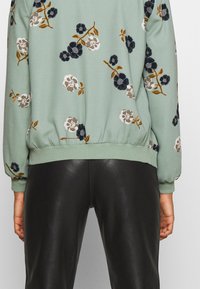 Grüner Sweatshirt mit langen Ärmeln, elastischem Saum und mehrfarbigen Blumenmustern, kombiniert mit schwarzen Lederhosen.