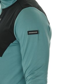 Langärmliges Sportshirt mit einem türkisfarbenen Körper, schwarzem oberen Paneel, strukturiertem Stoff und einem Markenlabel am Ärmel.