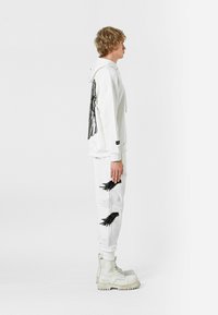 Sudadera blanca con bordado de alas negras en la parte posterior, combinada con pantalones de chándal blancos que presentan motivos de alas, y botas blancas gruesas.