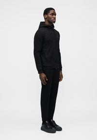 Melns sporta tērps no gludas auduma, kas sastāv no kapuces džempera ar rāvējslēdzēju un sašaurinātām biksēm, papildināts ar melniem sporta apaviem.