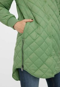 Giacca verde trapuntata con una tasca con zip, chiusura a bottone e polsini a coste. Texture morbida, design di media lunghezza e orlo arrotondato.
