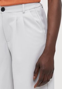 Hellgraue maßgeschneiderte Hose mit sichtbarem Bund, Falten und schwarzem Knopfdetail. Glatte Textur und gerade Beinform.