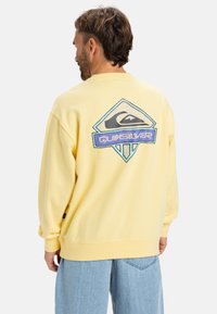 Yellow sweatshirt featuring a large graphic logo on the back, ribbed cuffs, and hem, paired with light blue jeans. Soft texture.