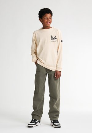 Beige T-shirt met lange mouwen en een grafisch ontwerp, gecombineerd met olijfgroene broek en zwarte sneakers met witte accenten.