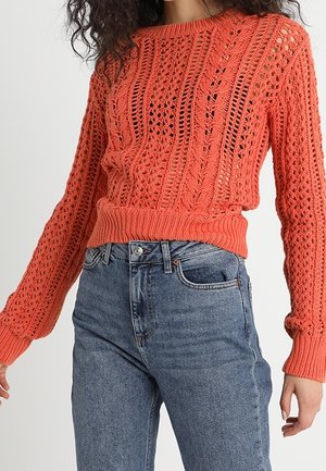 Femme portant un pull en maille orange corail avec des motifs torsadés et un jean en denim bleu taille haute sur un fond uni.