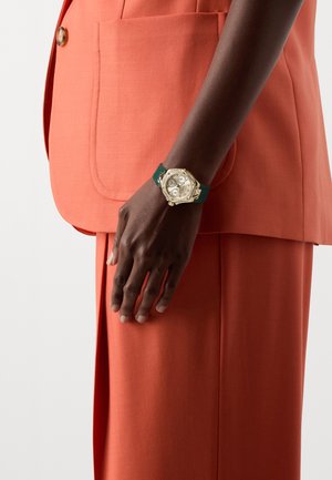 Montre octogonale dorée avec des cadrans blancs, bracelet en caoutchouc vert, portée sur une main contre un costume orange, mettant en avant le contraste de couleur et le design.