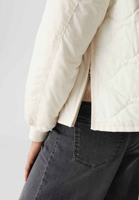 Veste matelassée beige clair avec des poignets côtelés, fermeture éclair et coupe décontractée, associée à un jean taille haute gris foncé.