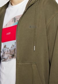 Sudadera con capucha y cremallera en verde oliva, con cordones en la capucha, que presenta un logotipo en el pecho y una camiseta gráfica blanca debajo, con palmeras.