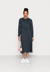 Vestido midi floral em preto com um padrão colorido. Mangas longas e cintura ajustada, combinado com ténis brancos e uma pequena mala rosa.