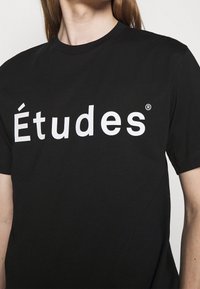 Personne portant un t-shirt noir avec le mot "Études" imprimé en grandes lettres blanches sur la poitrine.