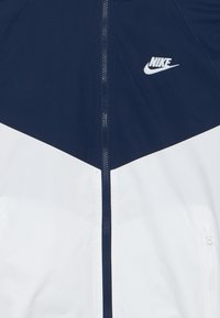 Jakna Nike z zgornjim delom v mornarsko modri barvi in spodnjim v belem, s popolnim zadrgo in izstopajočim logotipom na prsih. Gladek teksturni videz.