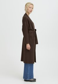 Donna con capelli biondi che indossa un lungo cappotto marrone con cintura, jeans ampi blu e scarpe nere a pianta bassa, in piedi di profilo su uno sfondo bianco.