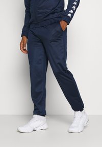 Tengerészi melegítőnadrág sima textúrával, rugalmas derékkal és logós díszítéssel. Fehér sportcipővel párosítva, amely strukturált mintázattal rendelkezik.