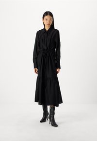 Musta midi-pitkä mekko, jossa on sidottava vyötärö, teksturoitu kangas, pitkät hihat ja kerrostettu helma. Yhdistetty mustiin kärjellisiin nilkkureihin.