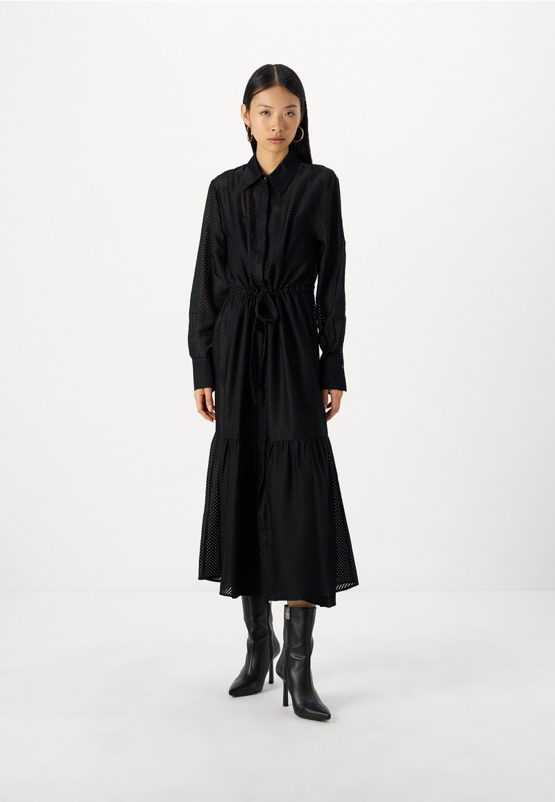 Musta midi-pitkä mekko, jossa on sidottava vyötärö, teksturoitu kangas, pitkät hihat ja kerrostettu helma. Yhdistetty mustiin kärjellisiin nilkkureihin.