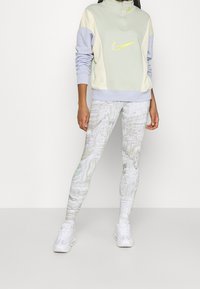 Person klädd i en pastellfärgad Nike-sweatshirt med färgblockering, vita mönstrade leggings och vita sneakers som står mot en enfärgad bakgrund.