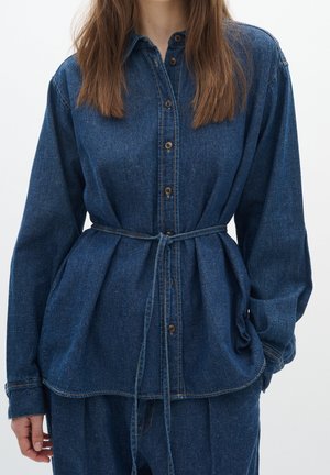 Vrouw draagt een blauwe denim blouse met knopen en een ceintuur in de taille, gecombineerd met bijpassende denim broek, staand tegen een effen witte achtergrond.