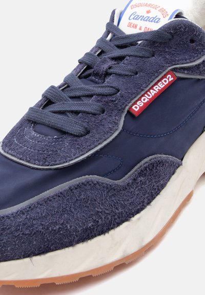 Gros plan sur une basket en suède et tissu bleu marine avec des lacets foncés, une semelle blanche, une étiquette rouge DSQUARED2 et une languette portant la marque Canada.
