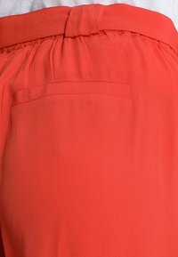 Korálově zbarvená, lehká textilie s hladkou strukturou, opatřená nařaseným pasem a malou kapsou na zadní straně.