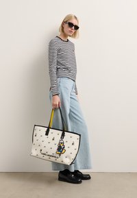White tote bag with black trim, featuring a cartoon character and bee illustrations. Model wears a black-and-white striped shirt and blue jeans.