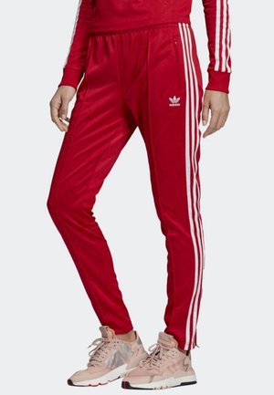 Osoba ubrana w czerwone spodnie dresowe Adidas z białymi paskami po bokach i beżowe trampki, stoi ze skrzyżowanymi nogami.