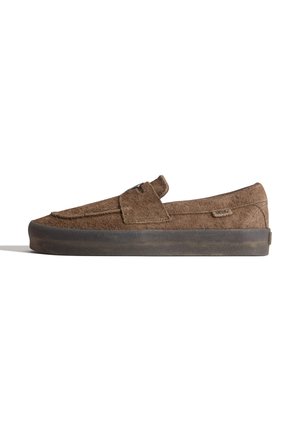 Mocassin en daim marron avec une semelle épaisse en caoutchouc foncé, doté d'une bride sur le dessus et d'une petite étiquette de marque en tissu sur le côté.