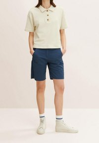 Jasno-beżowa koszulka polo z trzema brązowymi guzikami, zestawiona z ciemnoniebieskimi szortami. Model nosi białe trampki za kostkę i w paski skarpetki.