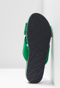 Tongs vertes avec une semelle en caoutchouc noir texturé, des sangles rembourrées et un matériau supérieur lisse. La semelle présente un motif de semelle en zigzag.