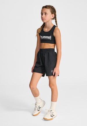 Jong meisje met gevlochten haar, gekleed in een zwart sportbeha en korte broek, witte sokken en sneakers, staand tegen een effen achtergrond.