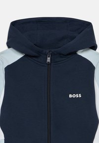 Navy zilā kapuce ar rāvējslēdzēju, kontrastējošiem gaiši zilā plecu akcentiem un balto "BOSS" logo uz krūts. Mīksta tekstūra.