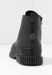 Botas negras hasta el tobillo hechas de cuero suave, con suela estriada, cierre de cremallera y un logo circular texturizado en la parte trasera.