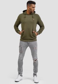 Sudadera con textura en verde oliva, con bolsillos delanteros y cordón, combinada con vaqueros rotos de color gris claro y zapatillas blancas.