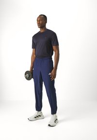 Marineblaues T-Shirt und passende Jogginghosen aus leichtem Material, in einer Hand eine Hantel haltend, weiße Schuhe mit schwarzen Akzenten.