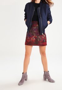 Femme portant une veste bomber navy, un haut noir, une mini-jupe à motifs rouge et violet, et des bottines en daim grises, debout les mains dans les poches.
