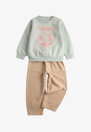 Sudadera verde claro con gráfica de pez koi rosa y texto "¡QUÉDATE KOI!", combinada con pantalones beige texturizados con bolsillos frontales.